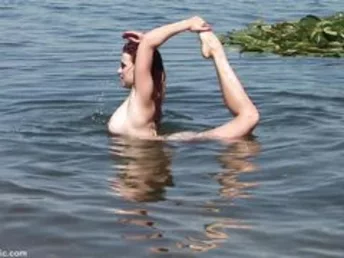 Smiling mermaid with red hair takes off the brassiere and performs flexing poses on the beach of large river.